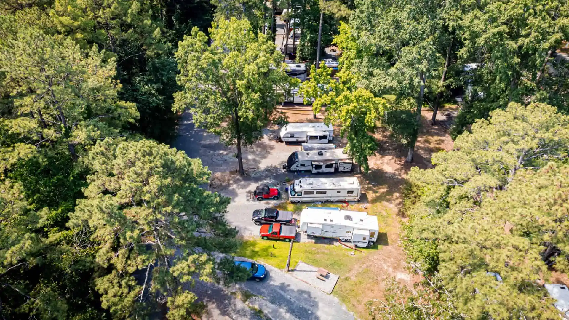 Aerial view of a campground with several parked RVs surrounded by tall green trees. A few cars are parked next to the RVs on gravel paths, and a small picnic area is visible. The scene is sunlit, showcasing a peaceful, natural setting.