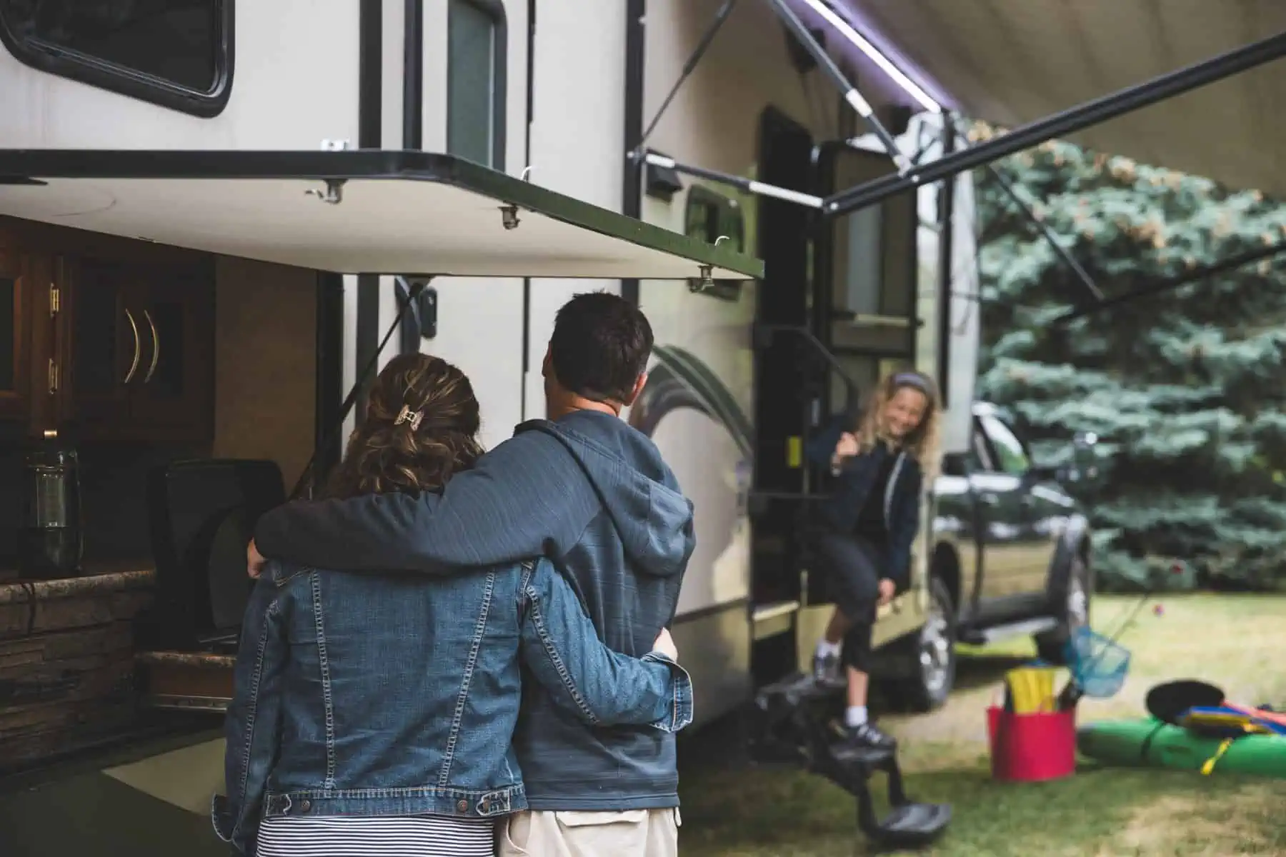 A couple with their backs to the camera stands together, looking at a woman seated in the doorway of an RV. There are trees and camping gear visible outside in a grassy area.