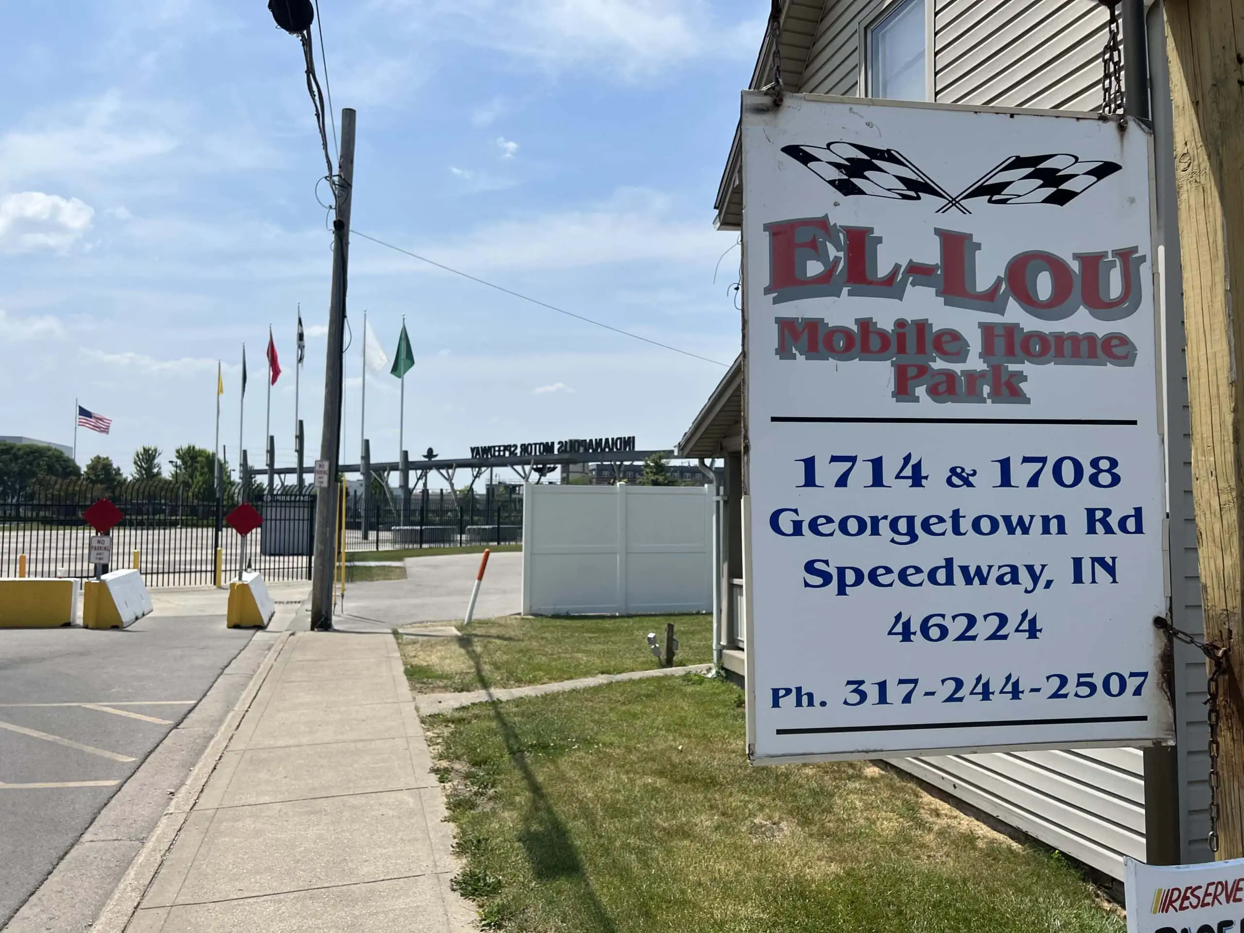Sign for El Lou Mobile Home Park with an address and phone number. In the background, an entrance gate with multiple flags and "Indianapolis Motor Speedway" signage. Sidewalks and street visible under a partly cloudy sky.